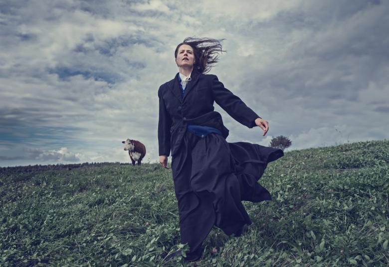 Kuvassa on pellolla rinteessä Emma Klingenberg mustassa pitkässä hameessa. Taaempana on ruskea-valkoinen lehmä.  Taustalla on harmaan sinistä pilvitaivasta. 