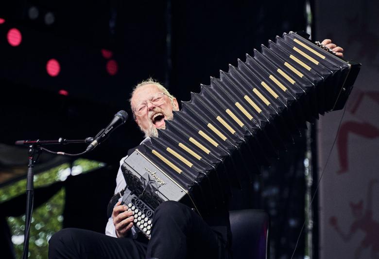 Kuvassa on Antti Paalanen, joka soittaa ja venyttää keskellä kuvaa hanurin palkeita täydessä mitassa. Taustalla on tummaa näyttämöä.