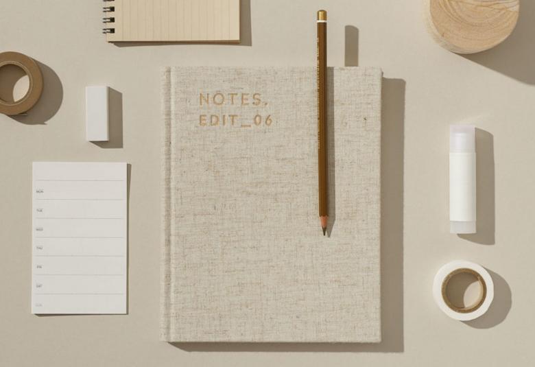 Kuvassa on vaalean harmahtavan ruskealla taustalla muistikirja, pieniä muistilehtiö,  kynä, teippirulla, vaalea pyyhekumi ja liimapuikko.