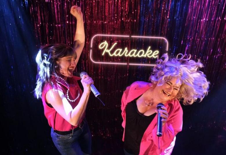 Kuvassa on vasemmalla vasen käsi pystyssä Annina Rubistein punaisessa puserossa ja farkuissa sekä hiuksissaan vaaleaa tekokarvaa. Oikealla on Johanna Heimonen eteenpäin vaaleat hiukset hulmuten punaisessa puserossa ja mustassa topissa. Molemmat laulavat mikit kädessään. Taustalla on karaoke-kyltti.