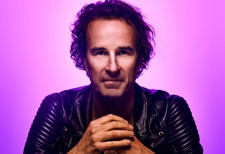 Kuvassa on Lauri Schreck ja kuvan tausta on violetti.  Schreckillä on tummat kiharaiset hiukset ja musta nahkatakki päällään.  Hänellä on kädet toistensa päällä edessään.