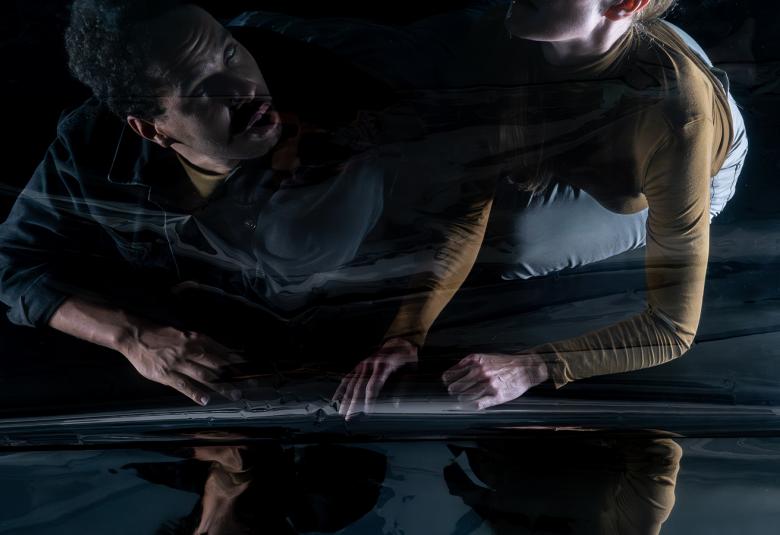 Kuvassa ovat vasemmalla mies ja oikealla nainen, jotka ovat heijastavassa pinnassa puolittain makuuasennossa. Kuvan sävy on tumma.