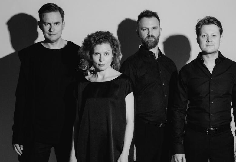 Kuvassa on hiukan taaempana kolme miestä ja hiukan heidän edessään naishenkilö.  Heillä kaikilla on mustat vaatteet päällään ja he seisovat. Taustalla näkyy heidän varjonssa.  Kuva on musta-valkoinen.