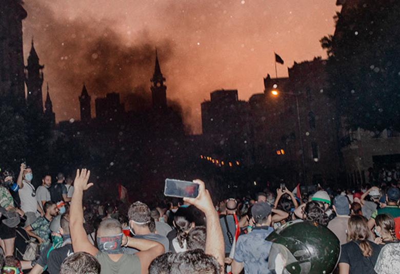 Kuvassa on alaosassa väkijoukkoa ja jotkut nostavat käsiää ja jotkut kuvaavat kännykällä edessä olevaa savuavaa kaupunkia. Sieltä nousee ruskeata savua.