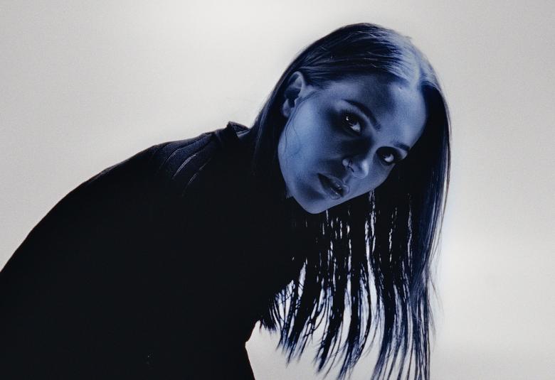 Kuvassa on Sanni kumartuneena oikealle puolivartalokuvassa mustassa puserossa. Hiukset ovat pitkät.  Kuva on musta-valkoinen.