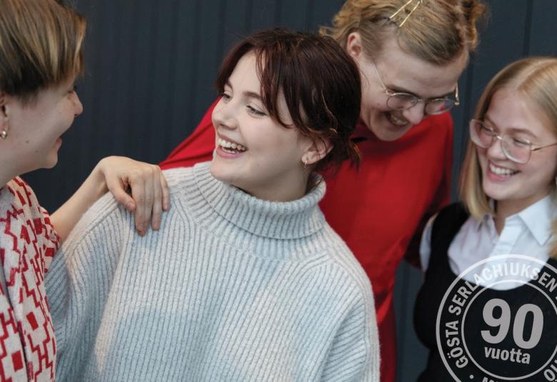 Kuvassa on vasemmalla nainen keskustelemassa nuoren naisen kanssa, jolla on vaalea villapaita. Hänen takanaan on ylempänä nainen katse alhaalla keskustelmassa oikealla puolella olevan nuoren vaaleatukkaisen naisen kanssa.