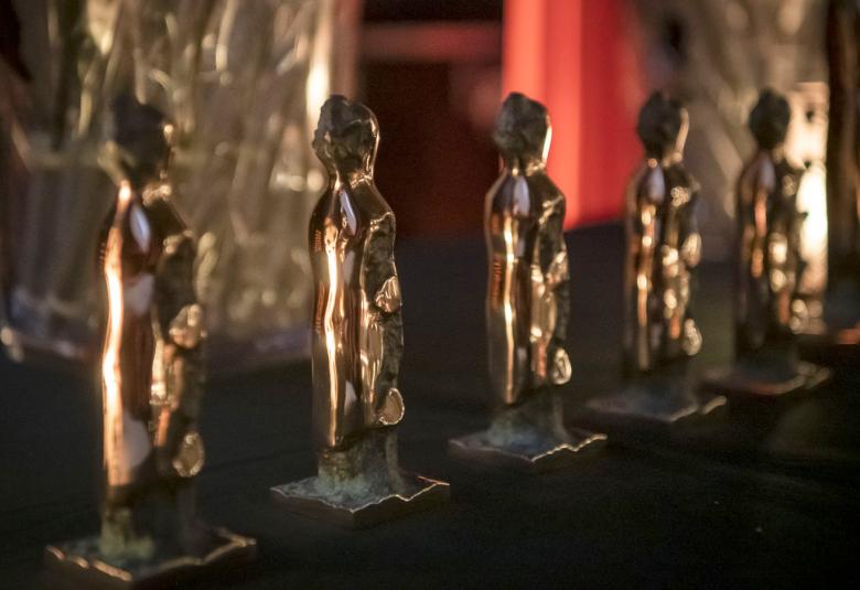 Kuvassa on vinosti viisi kultaista palkintopatsasta. Kuvan sävy on tumma. 