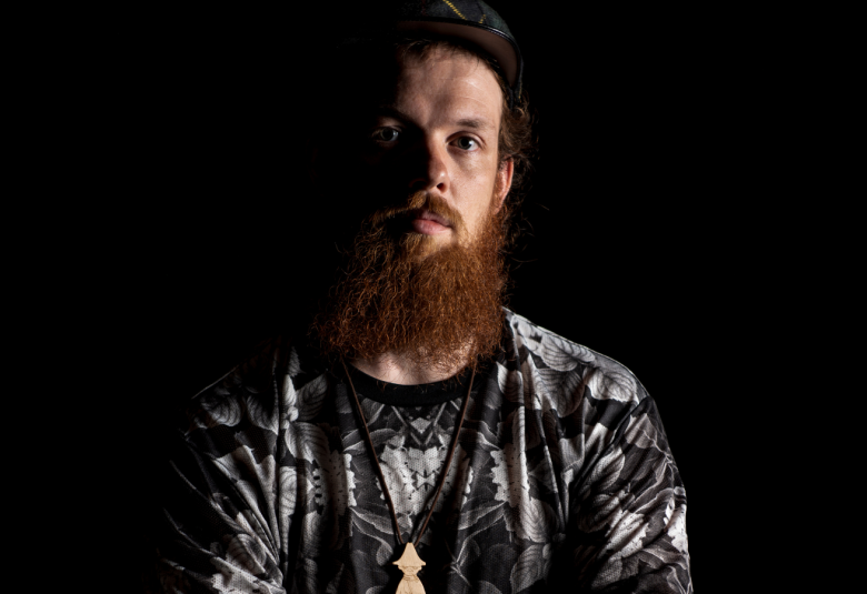 Kuvassa seisoo Taneli Törmä kädet poikittäin vatsan päällä.   Hänellä on mustan kiiltävä paita ja punertava parta.  Kuvan tausta on tumma.