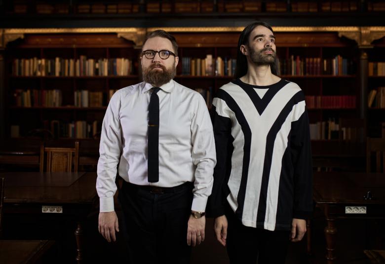 Kuvassa ovat seisomassa valkoisessa paidassa ja mustassa solmiossa Juuso Koponen ja musta-valkoisessa isoja v-kuvioisessa villapaideassa Jonatan Hildén seisomassa. Hilden katsoo etuviistoon oikealle. Takana on kirjahyllyä ja tausta on tummahko.