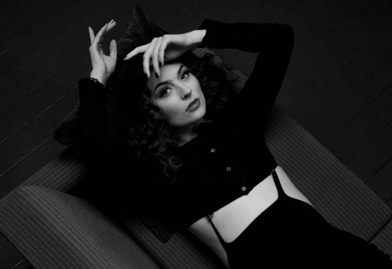 Kuvassa on BEHM puoliksi makuulla. Hänellä on musta, lyhyt pusero päällään ja vasen käsi otsalla. Toinen käsi on pään päällä. Kuva on musta-valkoinen.