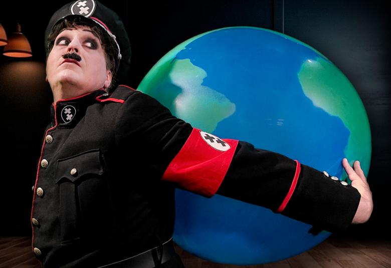 Kuvassa on Juha Pulli Hynkelinäja hänellä on musta uniformu päällään ja siinä on punaiset raidat reunoissa sekä punainen nauha kädessään. Päässä on musta koppalakki, jossa on valkoiset ja punaiset raidat.  Takanaan hän pitää ilmapalloa, jossa on maapallon kuva.