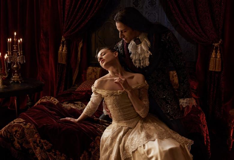 Kuvassa on Draculaa esittävä tanssija ylhäisöasussa ja hän on oikealla kumartuneena nuoren naisen puoleen, joka on vaaleassa pitkässä mekossa ja nojaa taaksepäin vuoteen päädyssä. Huoneessa on tumman burgundin väriset samettiverhot ja saman sävyistä peittoa sekä kalusteita.