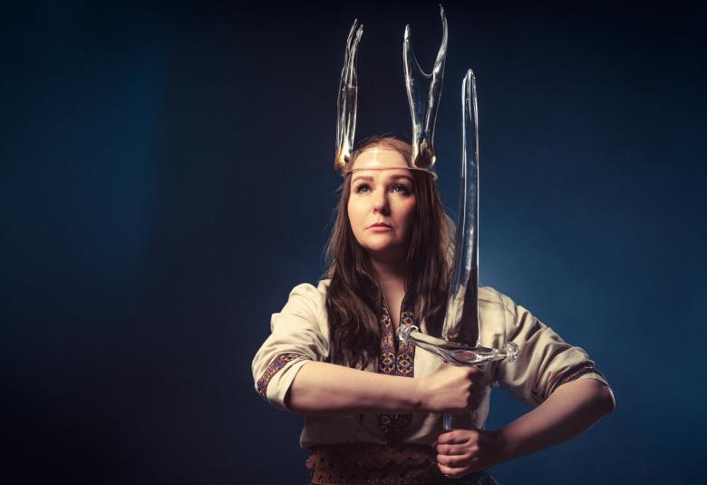 Kuvassa on Sini Majuri vaaleassa puserossa, päässään lasikruunu ja kädessään pystyssä oleva lasimiekka. Tausta on tumma.