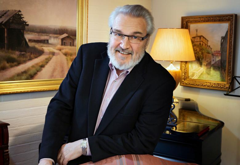 Kuvassa on Jaakko Ryhänen istumassa ja hän nojaa pöytään.  Kuva on puolivartalokuva. Taustalla näkyy kaksi taulua ja pöytälamppu. Ryhäsellä on musta takki ja valkoinen paita päällään.