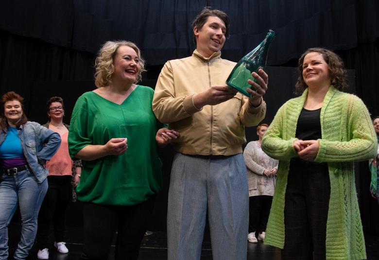 Kuvassa on etualalla vasemmalla vaaleatukkainen nainen vihreässä puserossa.  Keskellä on mies kerllertävässä pusakassa ja hän pitää kädessään isoa vihreää kuohuviinipuolloa.  Okealla puolella on nainen, jolla on vaalean vihreä pitkä villatakki.  Taustalla näkyy naisia farkuissa ja erilaisissa puseroissa.