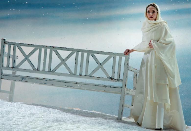  Kuvassa on taustalla vaalean sini-valkoista taivasta.  Edessä on vinosti lumihanketta ja sen päällä on harmahtava penkki, jossa on selustassa pystypuita ja ristissä olevia puita.  Vieressä on oikealla Tatjana valkoisessa, pitkässä puvussa.    