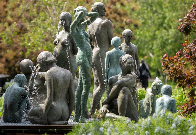 Kuvassa on ryhmä patsaita naisista ja lapsista selin seisomassa ja istumassa vihreiden lehtipuiden ja pensaiden keskellä suihkulähteen äärellä. Patsaat ovat vihreän harmaita.