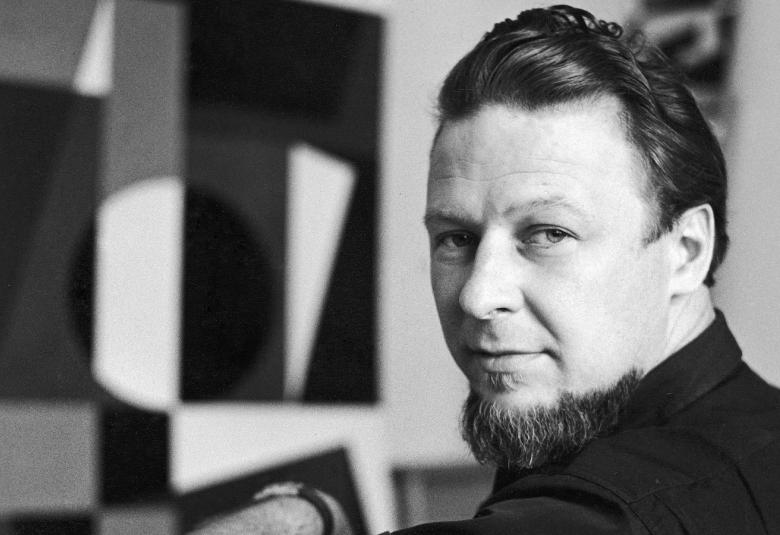 Kuvassa on Lars-Gunnar Nordström, joka istuu ja katsoo olkapäänsä yli. Taustalla näkyy taiteilijan teos epätarkasti. Kuva on musta-valkoinen.