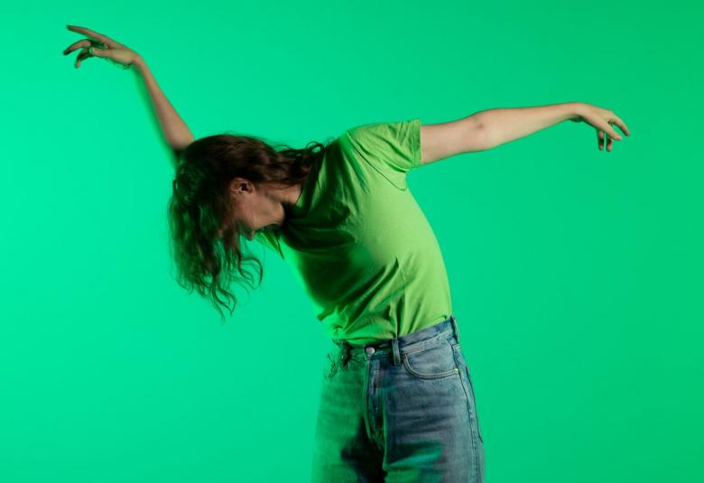 Kuvassa on vihreällä taustalla Liisa Pentti tanssimassa puolivartalokuvassa. Hänellä on pää alaspäin ja oikea käsi ojennettuna ylöspäin ja vasen käsi ojennettuna sivulle. Pentillä on päällään vihreä t-paita ja siniset farkut.