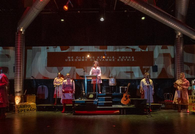 Kuvassa on tummahko näyttämö ja siellä on takana vaaleata seinää sekä pylväillä rajattua rakennelmaa.  Näyttämöllä on useita henkilöitä seisomassa vaaleahkoissa vaatteissa. 