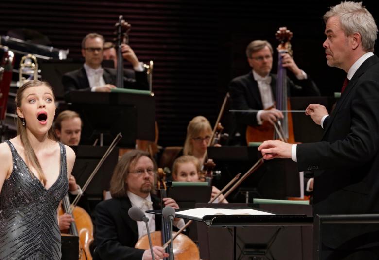 Kuvassa on laulamassa Johanna Wallroth vasemmalla mustassa, kiiltävässä hihattomassa puvussa ja taustalla on orkesteri.  Oikealla selin on Hannu Lintu johtamassa orkesteria.