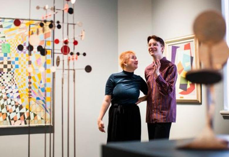 Kuvassa on vasemmalla keltaisen sävyinen pikkuruutuihin ja geometrisiin kuvioihin perustuva taideteos ja sen edessä kapeisiin teräsputkiin kiinnitettyihin pieniin pyöreisiin metallilätkiin perustuva teos.  Oikealla on nainen sinisessä puserossa ja nuorehko mies violetissa puserossa ihailemassa teosta.