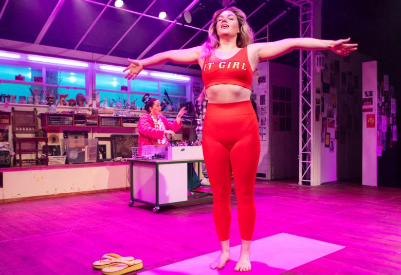 Kuvassa on nainen keskellä pinkkiä lattiaa ja oikealla takana on nainen kirjoituspöydän äärellä. Taustalla näkyy salia pöytineen.