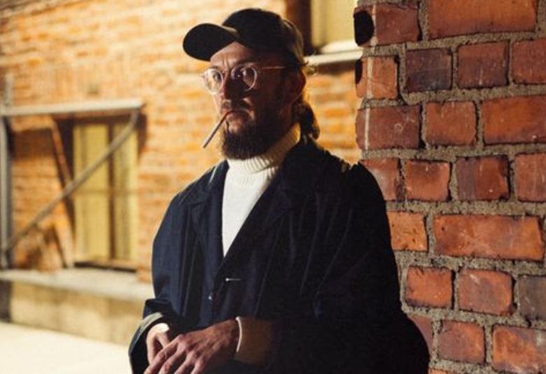 Kuvassa on Samuli Putro puolivartalokuvassa ja taustalla näkyy varaston ruskeaa tiiliseinää. Hän seisoo tummansinisessä pusakassa ja hänellä on lippalakki, silmälasit ja tupakka roikkumassa huulesta. 