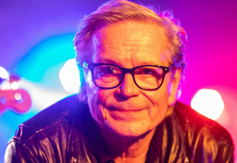 Kuvassa on Kake Randelin kasvokuvassa.  Hän katsoo suoraan eteenpäin ja hänellä on silmälasit sekä musta nahkatakki.  Taustalta tulee violettia valoa oikealle poskelle.  Taustalla on turkooseja ja vielettejä valoja.