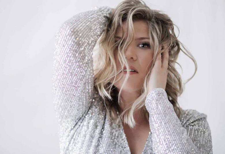 Kuvassa on Katri Ylander puolivartalokuvassa ja hänellä on vaaleat kiharaiset hiukset, joita hän pitää käsillään. Oikea käsi on ylempänä ja vasen käsi on posken päällä. Ylanderilla on valkoinen pusero päällään ja tausta on vaalean harmahtava.