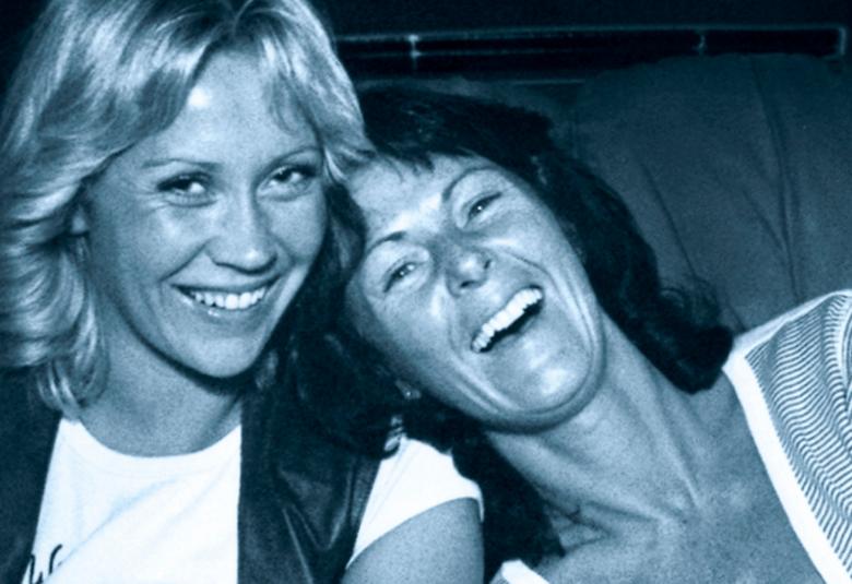 Kuvassa ovat Agneta Fältskog ja Ann-Frid Lyngstad kasvokuvassa.  Vasemmalla on vaaleatukkainen Fältskog ja häneen nojaa tummatukkainen Lyngstad. He molemmat nauravat.