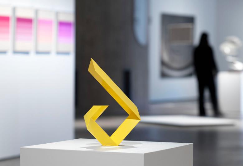  Kuvassa on etualalla näyttelytilaa valkoisella korokkeella keltainen leveästä metallista taitettu alhaalla olevalla kaarella ylöspäin taittuva kuvio.