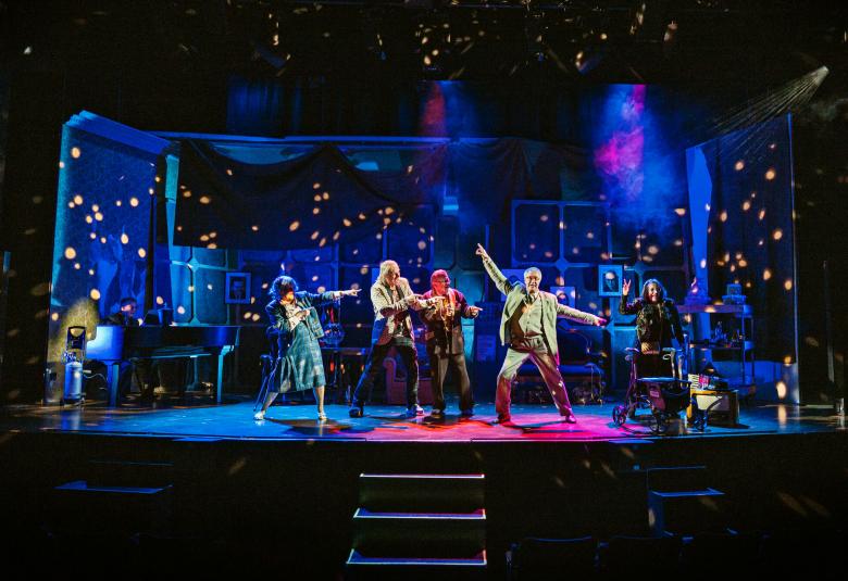 Kuvassa on tumman sinisävyinen näyttämö ja siinä on viisi näyttelijää vauhdikkaasti laulamassa ja liikehtimässä. Oikealla on mies vaalean ruskeassa puvussa ja kädet ojennettuina sivuille.