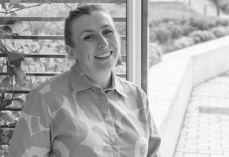 Kuvassa on Heidi Länsisalmi puolivartalokuvassa. Hänellä on Unikko-kuvioinen paitapusero päällään.  Vasemmalla on vaakaraudoitetussa portissa kiipeilevää viherkasvia ja oikealla nousevaa kivetystä ja vaaleaa kaidetta.