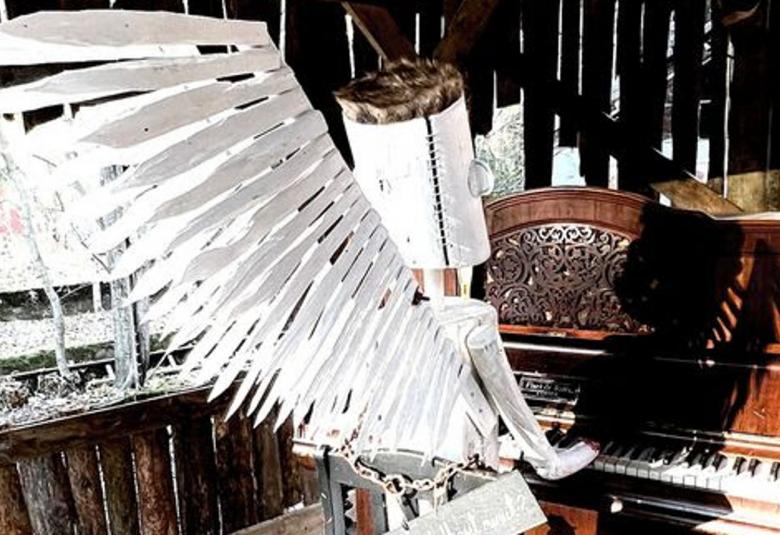 Kuvassa on tummassa raakapuusta rakennetussa tilassa vanhanaikaista pianoa soittamassa valkoisista siivistä tehty hahmo, jolla on torven muotoiset valkoiset käsivarret ja kädet soittamassa sekä valkoinen tötteröpää.