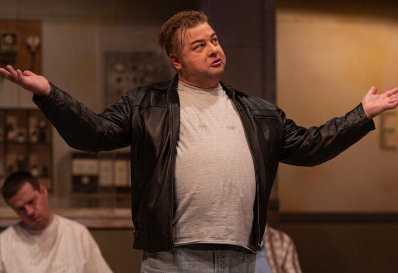 Kuvassa on Jan Mykkänen ja hän seisoo kädet nostettuna molemmille puolille. Hänellä on musta nahkatakki ja harmaa pusero päällään. Alempana istuu nuokkuen mies vaaleassa paidassa. 