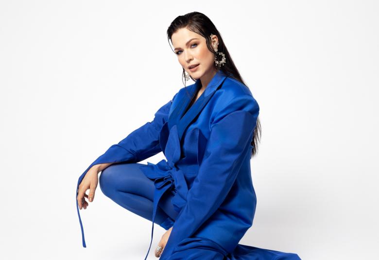 Kuvassa on Jenni Vartiainen sähkön sinisessä pitkässä, koko vartalon peittävässä puvussa ja siinä on laahus. Hän on kyykkyasennossa hieman sivuttain.  Tausta on valkoinen.