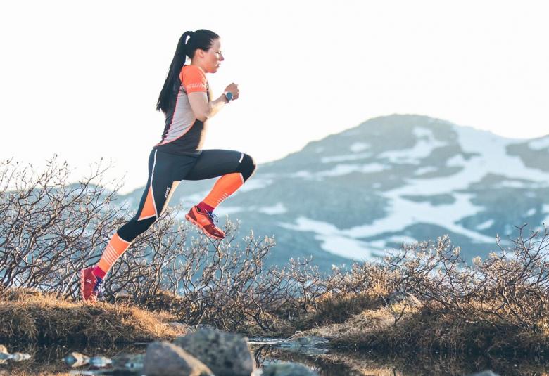Kuvassa on Krista Pärmäkoski hyppäämässä vuoristossa hyppäämässä ojan yli. Takana on pensaita. Taustalla näkyy vuoristoa. Hänellä on musta-oranssi treeniasu.