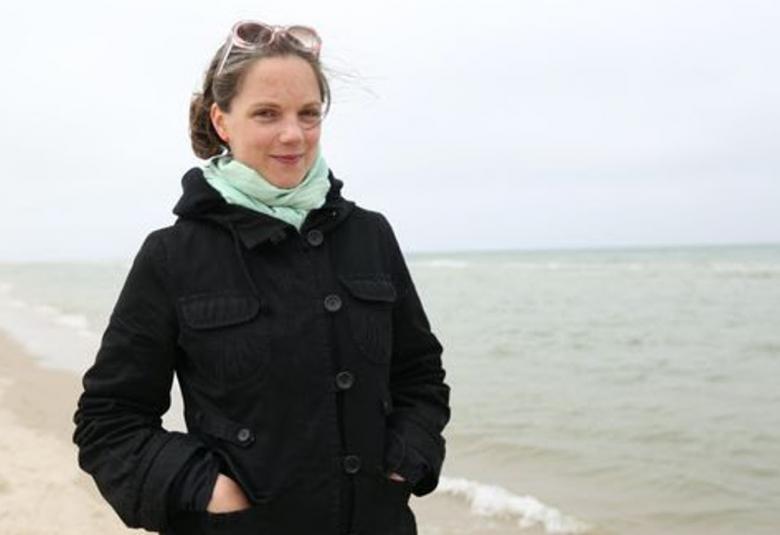 Kuvassa on Kukka Paavilainen mustassa takissa ja vaalean vihreässä huivissa. Hän seisoo kuvan vasemmalla puolella vaalealla hiekkarannalla, jossa näkyy merta.