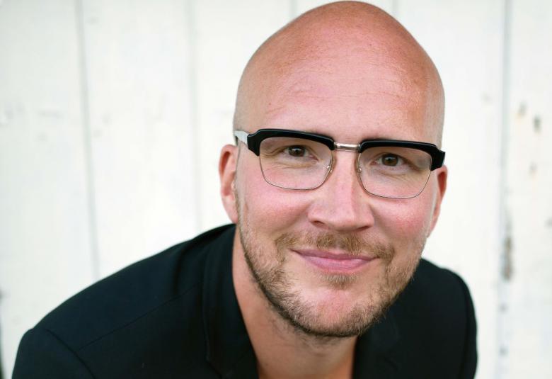 Kuvassa on Marzi Numanin kasvot ja hänellä on silmälasit eikä hiuksia.  Hänellä on musta pusero.  Taustalla on vaaleaa seinää.