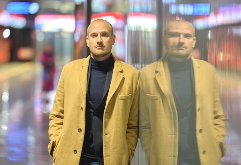 Kuvassa on Max Seeck keltaisessa takissa ja taustalla on himmennettynä Helsingin metroasemalaituria. Seeck on kahdennettu oikealle himmeämmin kuin vasemmalla eli on peilikuvassa.