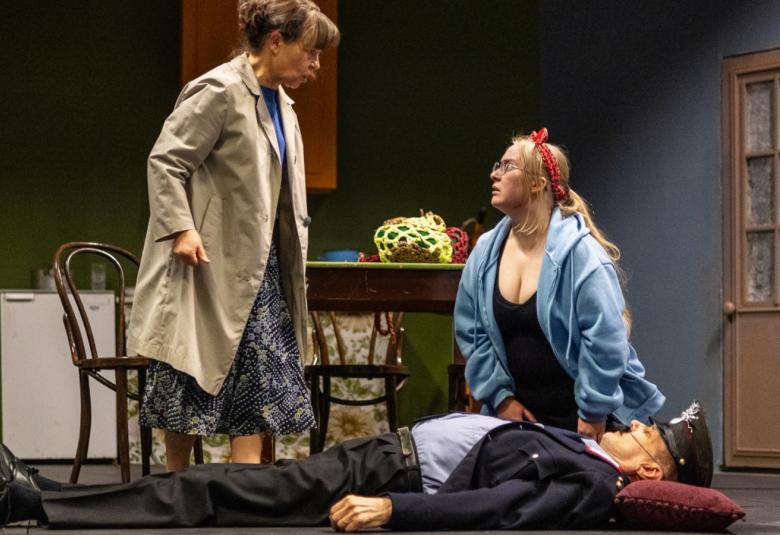 Kuvassa vaaleatakkinen nainen seisoo ja juttelee polvillaan olevan vaalean sinisen hupparisen naisen kanssa.  Edessä on mies tummas puvussa ja koppalakissa.