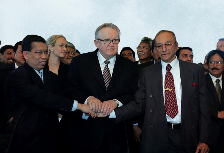 Kuvassa on keskellä Martti Ahtisaari ja hänen molemmilla puolillaan on mieshenkilöt ja he pitävät toisiaan kädestä kiinni. He ovat mustissa takeissa. Taustalla on paljon ihmisiä.