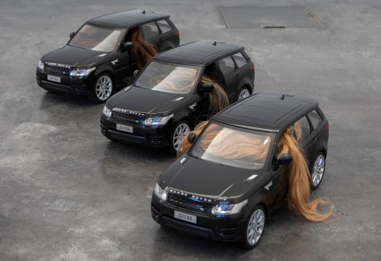 Kuvassa on harmaalla pohjalla tumman harmaita maastureita vinottain alaspäin. Alimpana olevan vasemmasta ikkunasta tulee pitkiä hiuksia ulos.