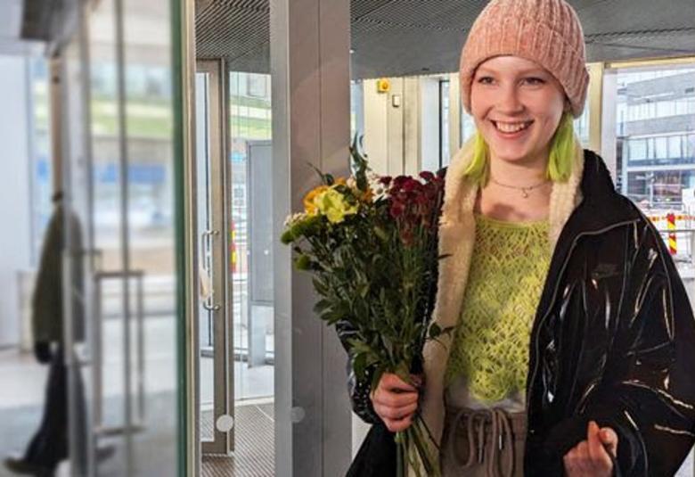 Kuvassa on Sonja, jolla on kiiltävän musta takki ja vaalean vihreä villapaita sekä vaalean ruskea pipo. Hänen oikeassa kädessään on kukkakimppu. Vasemmalla on lasisia ovia.