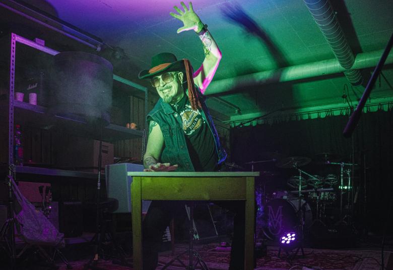 Kuvassa on mustassa lierihatussa ja sinisessä farkkuliivissä oleva mies rummuttamassa pöytäää oikealla kädellä ja vasen käsi on nostettu ilmaan.  Taustalla näkyy tummaa telttakatosta. 