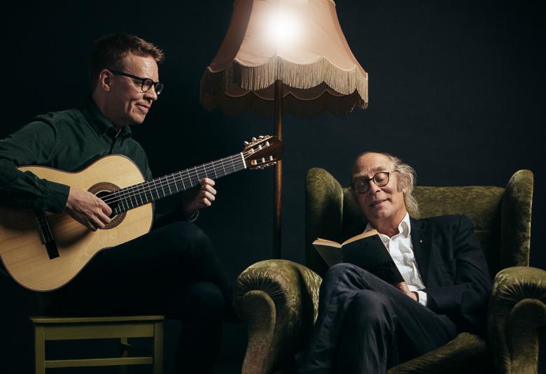 Kuvassa on vasemmalla Petri Kumela soittamassa kitaraa ja oikealla Vesa Vierikko istumassa isossa nojatuolissa. Taustalla on tummaa huonetta.