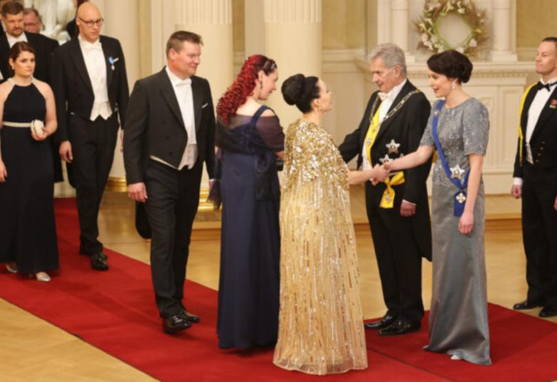 Kuvassa ovat oikealla Jenni Haukio ja Sauli Niinistö vastaanottamassa vieraita ja kättelemässä on melkein selin nainen punaisissa, kiharaisisssa hiuksissa ja mustassa pitkässä mekossa.  Hänen takanaan on mies mustassa frakissa.  Hänen takanaa näkyy pariskuntia tulossa kättelyyn punaisella matolla.  Lattia on ruskehtavaa parkettia. Presienttiparin takana on kaksi adjutanttia mustissa kadettiasuissa.