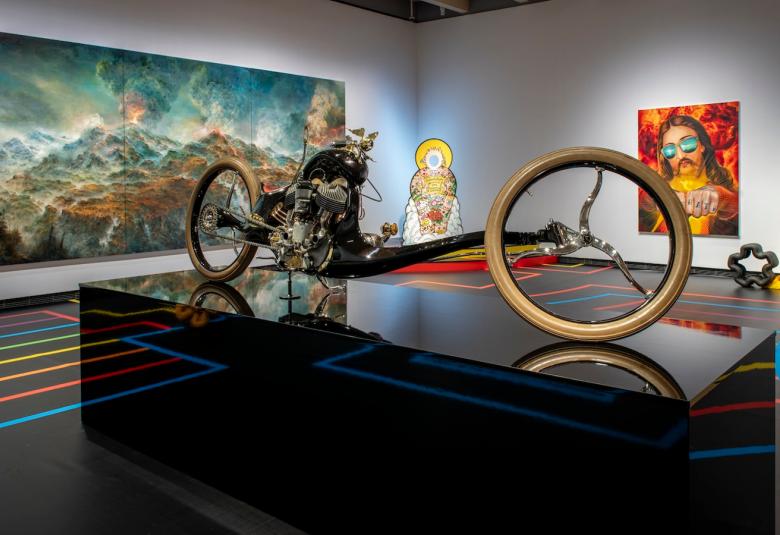 Kuvassa on rakennettu moottoripyörä, joka on lavan päällä. Etupyörä on pitkä rungon päässä.  Taustalla on tumman sinertävä ja ruskehtava vuoristomaisema. Toisella seinällä on pitkätukkaisen miehen kasvot ja hänellä on aurinkolasit. Tausta on oranssin punainen.