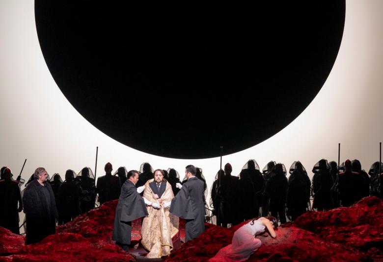 Kuvassa on suuri musta kuu vaalealla taustalla.  Alla on suuri joukko tummia hahmoja.  Edessä on punaista kangasta ja siinä seisoo vasemmalla mies mustassa kaavussa.  keskessä on mies sivuttain vaaleassa kaavussa olevan miehen vieressä. Oikealla makaa nainen maassa ja hänellä on vaalea puku.